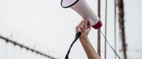 a person's arm raises a megaphone into the gray city sky