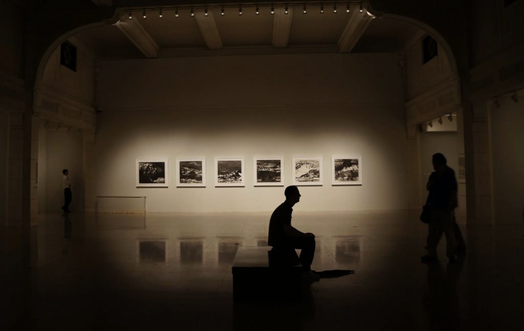 A silhouetted figure sits on a bench in an art gallery. 