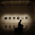 A silhouetted figure sits on a bench in an art gallery.
