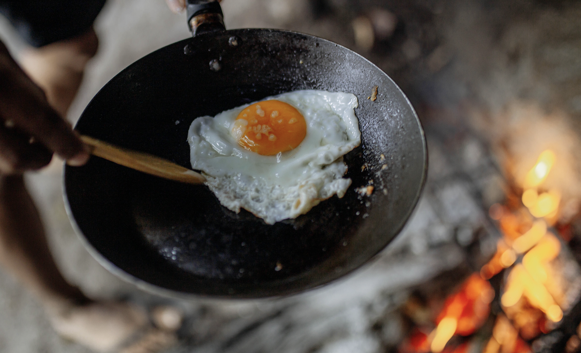 A person, barefoot, cooking an egg sunny-side up above an open flame