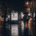 A couple walking through the city streets at night, under an umbrella in the rain