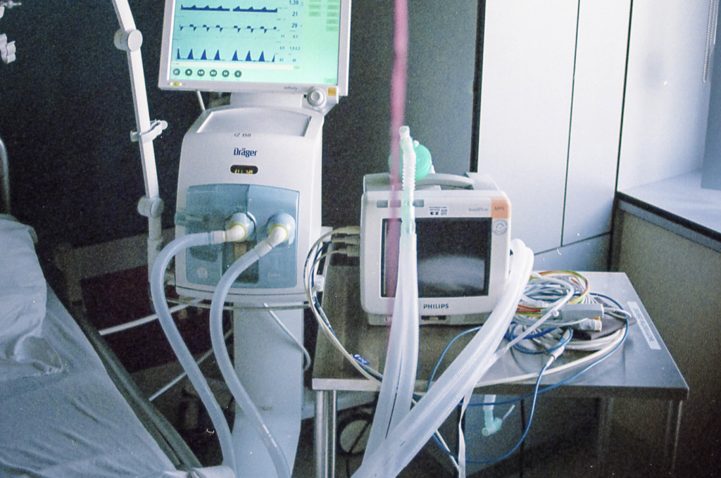 The edge of a hospital bed surrounded by tubes, wires and screens